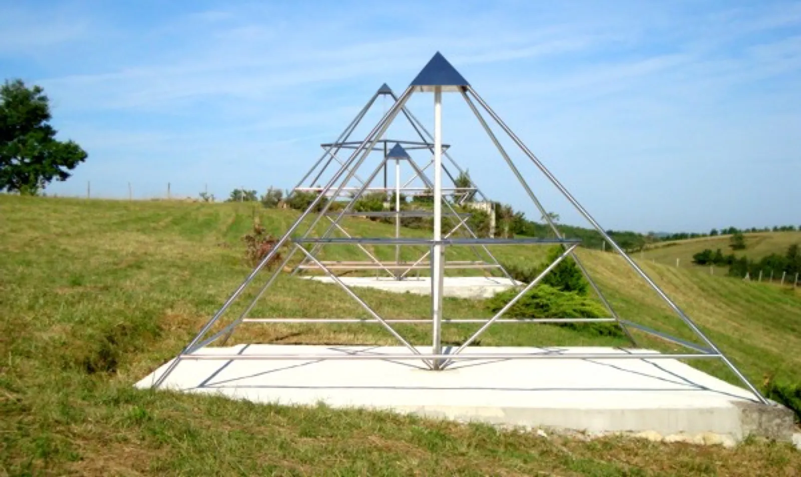 Große offene Energiepyramide aus Edelstahlrohren mit dunkler Spitze und Mittelachse auf einer frischen Betonplatte an einem Wiesenhang; dahinter stehen zwei weitere Pyramidengestelle, rechts Hügel und Baumreihen unter blauem Himmel.