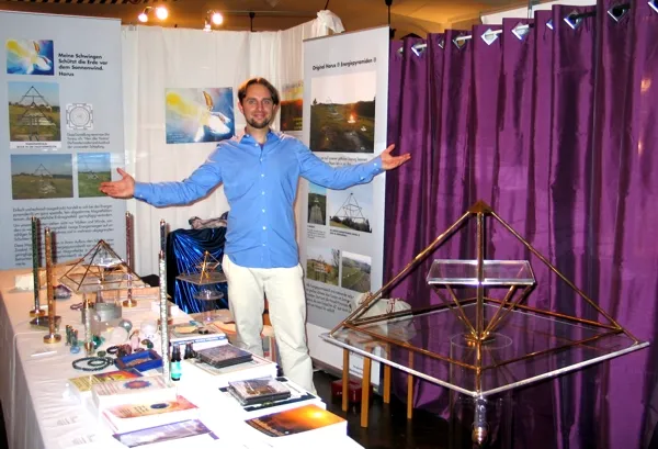 Andreas Grohmann im blauen Hemd mit ausgebreiteten Armen an seinem Messestand; links Tische mit goldenen Energiepyramiden, Kristallsäulen und Büchern, rechts eine große Horus-Energiepyramide vor violettem Vorhang.