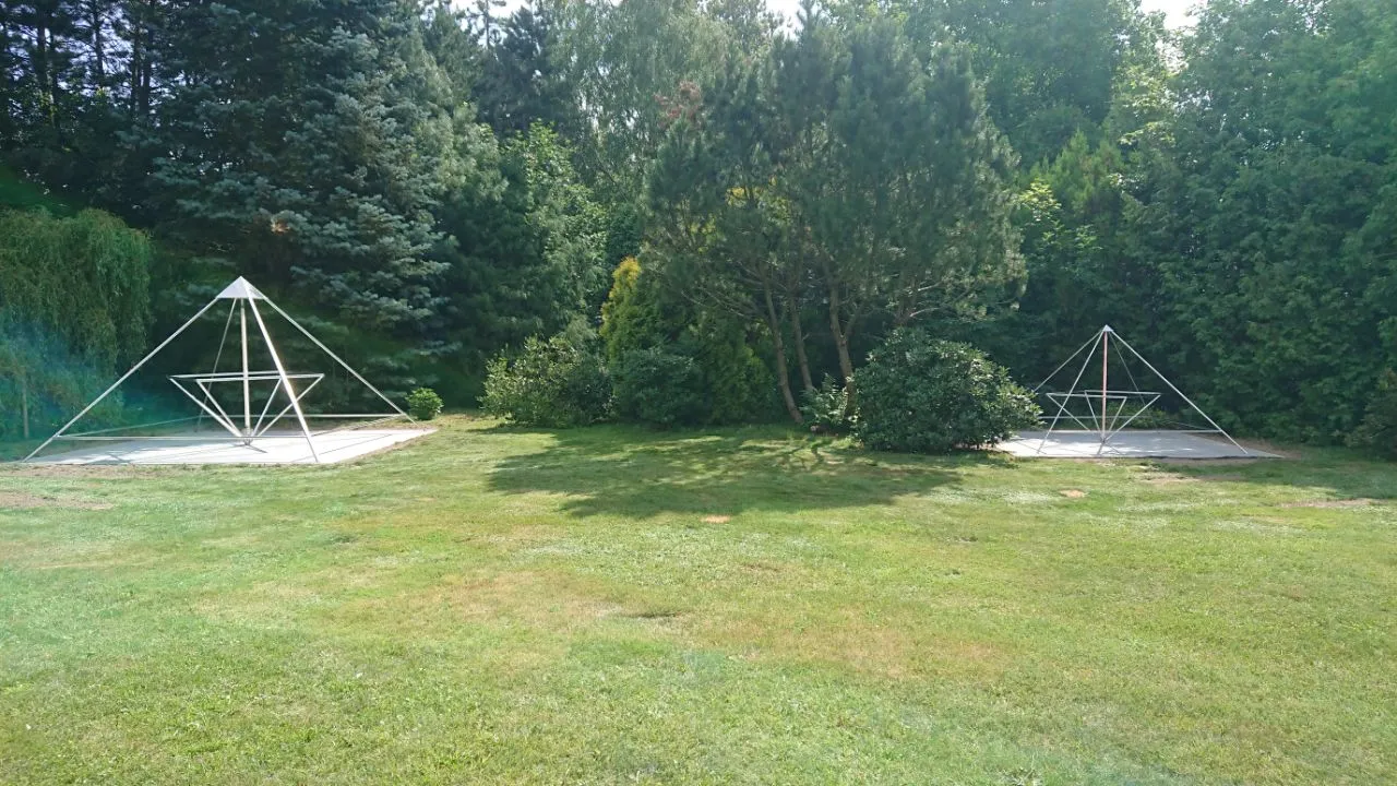 Eine große Gartenanlage in Polen mit zwei silbernen Metallrahmen-Energiepyramiden (Modell C und D) auf Betonsockeln, umgeben von Nadelbäumen.