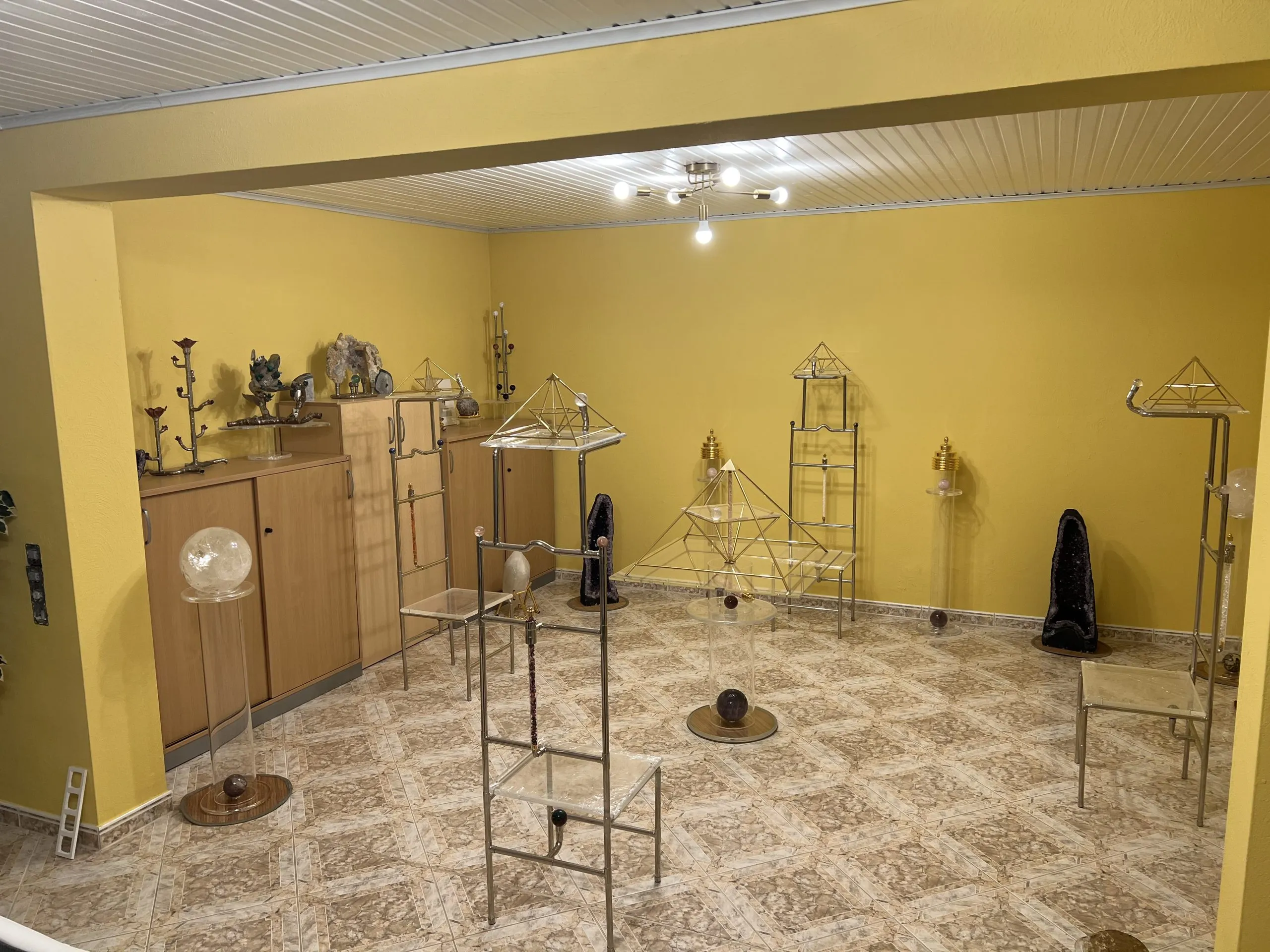 Renovierter Ausstellungsraum mit gold-gelben Wänden, in dem zahlreiche Energiepyramiden-Modelle auf Ständern, Kristallkugeln und dunkle Kunstobjekte präsentiert werden.