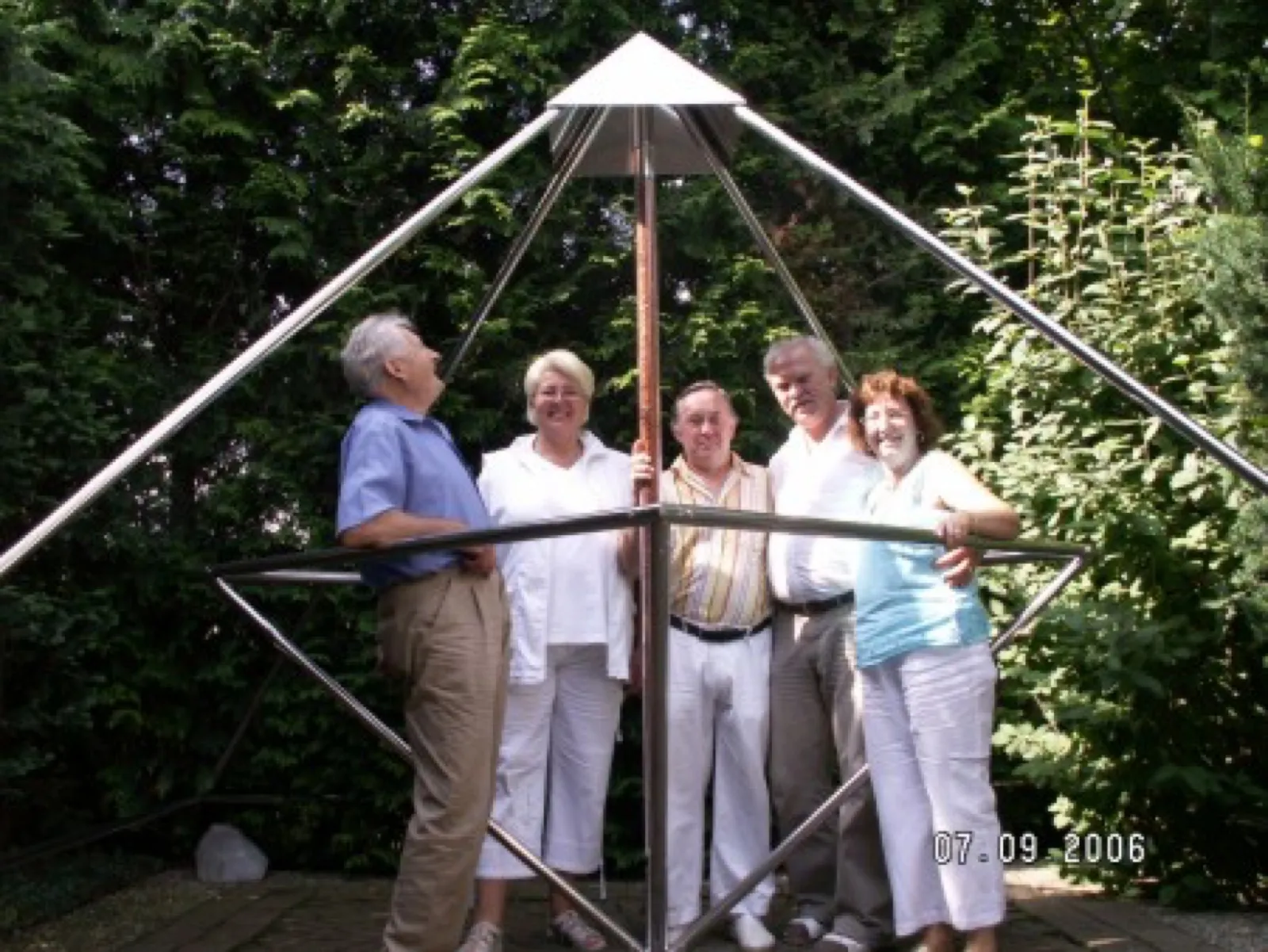 Fünf ältere Erwachsene stehen lächelnd unter einer großen offenen Edelstahlpyramide mit spitzem Deckblech in einem von hohen Hecken umgebenen Garten; einer der Männer blickt nach oben zur Spitze.