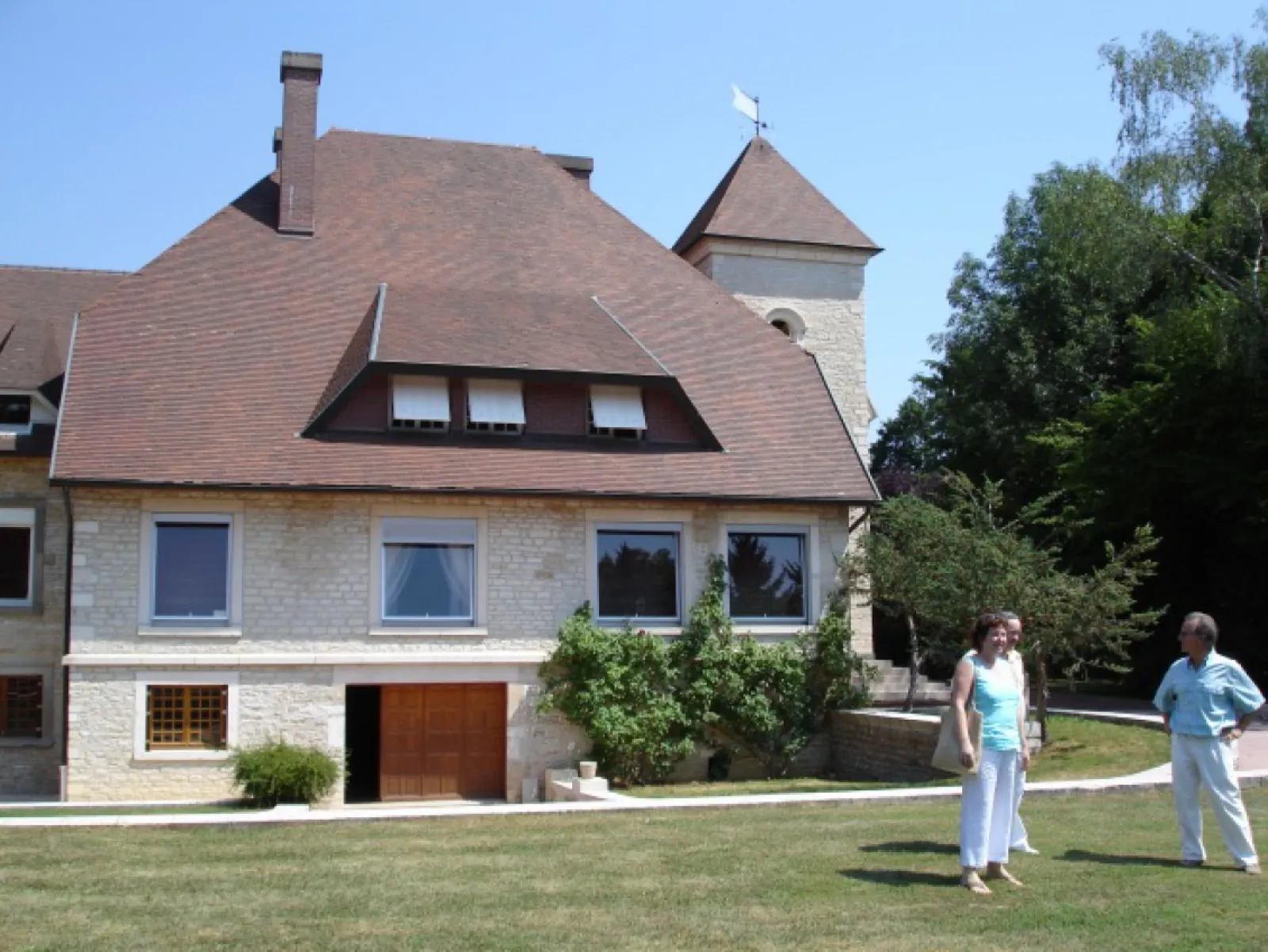Sommerliches Natursteinhaus mit steilem braunem Ziegeldach, Dachgauben und einem Turm mit Wetterfahne; auf dem Rasen davor stehen eine Frau in türkisfarbenem Oberteil, eine halb verdeckte Person und ein Mann in hellblauem Hemd.