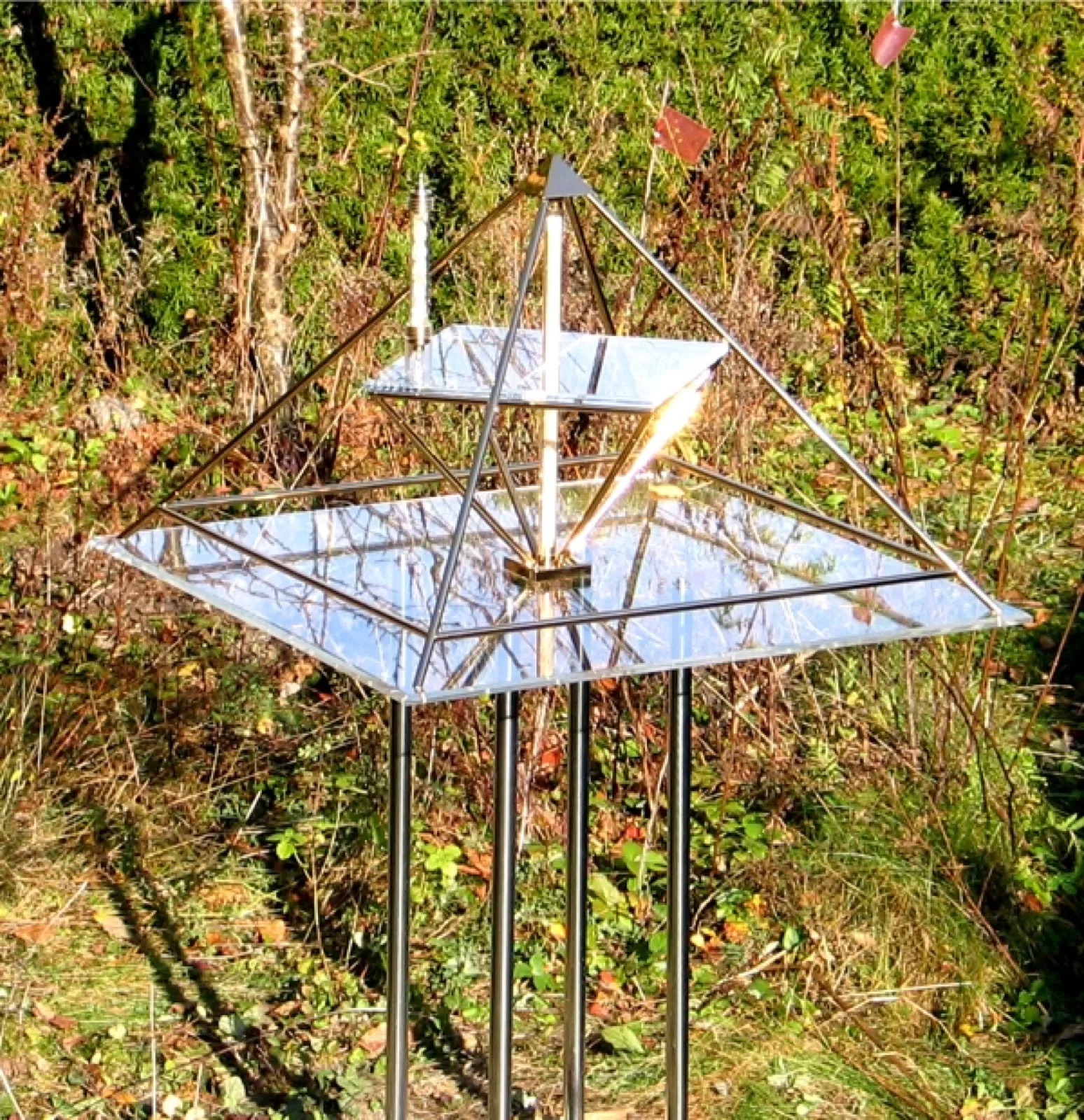 Energiepyramide mit goldfarben schimmernden Stäben, zwei spiegelnden Acrylglasplatten und senkrechter Mittelachse auf vier hohen Edelstahlbeinen im Freien, dahinter sonnenbeschienenes Buschwerk.