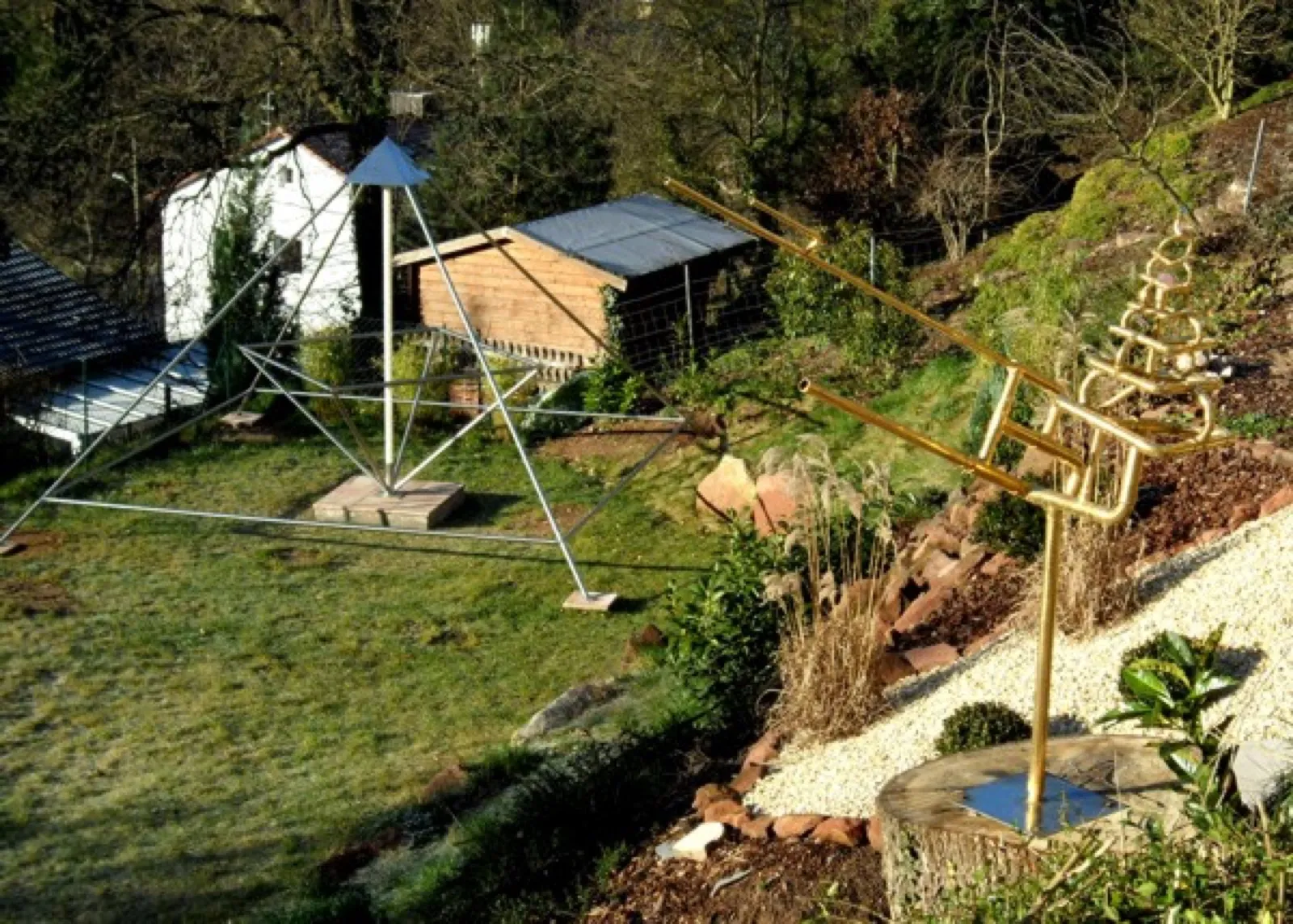 Hanggarten bei Landau in Wintersonne: Links steht eine offene Energiepyramide aus Metallstäben mit blaugrauer Spitze und innenliegendem Oktaeder auf einer Platte im Rasen, dahinter ein Holzschuppen und ein weißes Häuschen; rechts im Vordergrund die vergoldete Rohrskulptur Vidaya auf einem Baumstumpf neben einem Kiesbeet.
