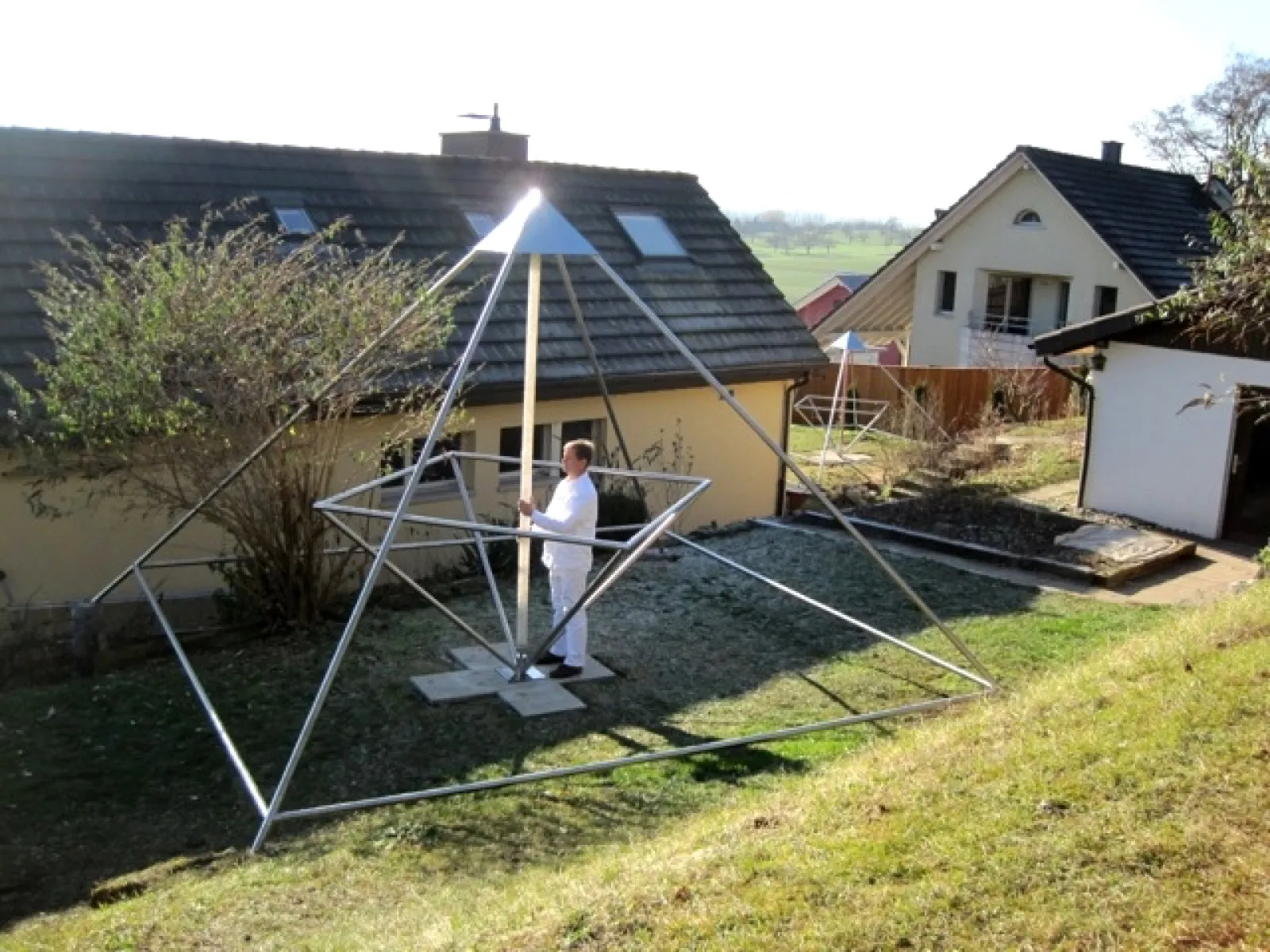 Ein weiß gekleideter Mann steht in einer großen offenen Energiepyramide aus Metallrohren mit hölzern wirkender Mittelachse in einem Hausgarten; im Hintergrund Wohnhäuser, ein Zaun und eine zweite, kleinere Pyramide.