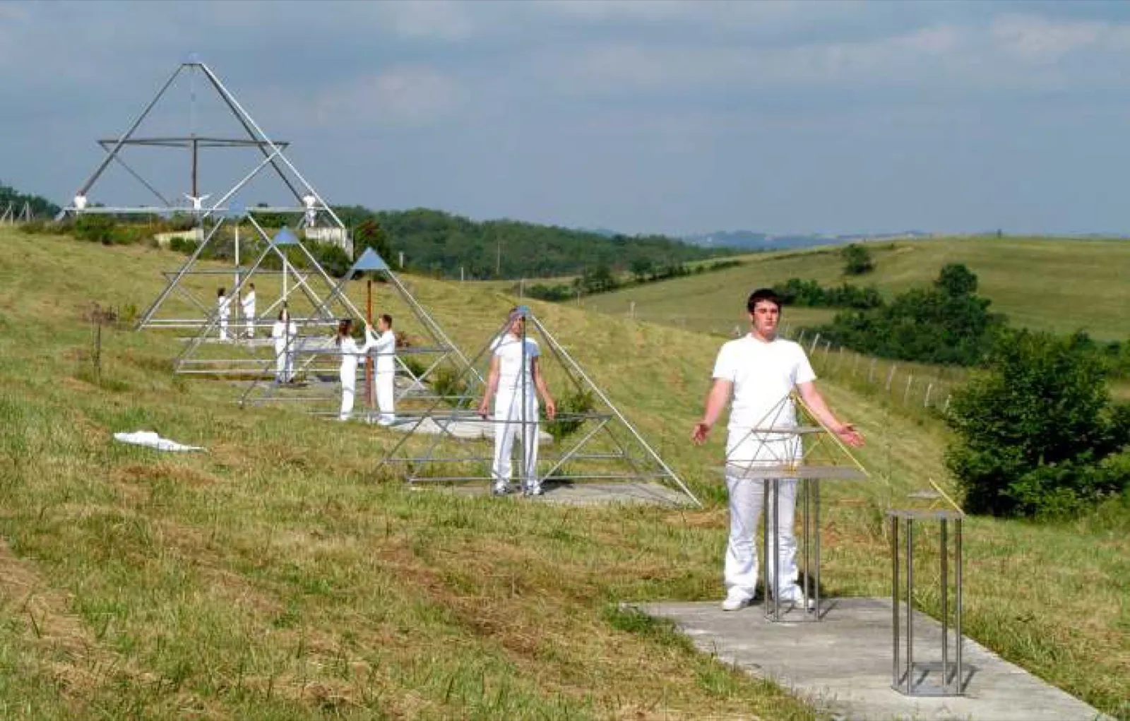 Weiß gekleidete Menschen stehen mit geöffneten Armen an Pulten und in mehreren offenen Pyramidengestellen, die gestaffelt einen grasbewachsenen Hang hinaufführen; ganz oben die größte Pyramide. Im Hintergrund hügelige Landschaft mit Hecken und Wald unter bewölktem Himmel.