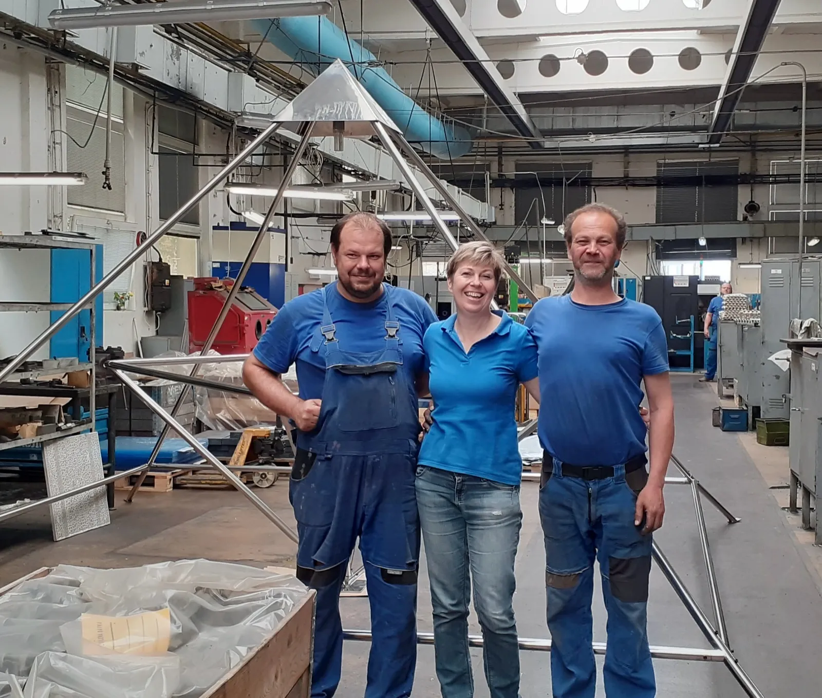 Zwei Männer und eine Frau in blauer Arbeitskleidung stehen lächelnd in einer Metallwerkstatt vor einer großen, noch nicht montierten Pyramide aus Edelstahlrohren mit glänzender Spitze; ringsum Regale, Maschinen und Deckenleitungen, hinten ein weiterer Arbeiter.