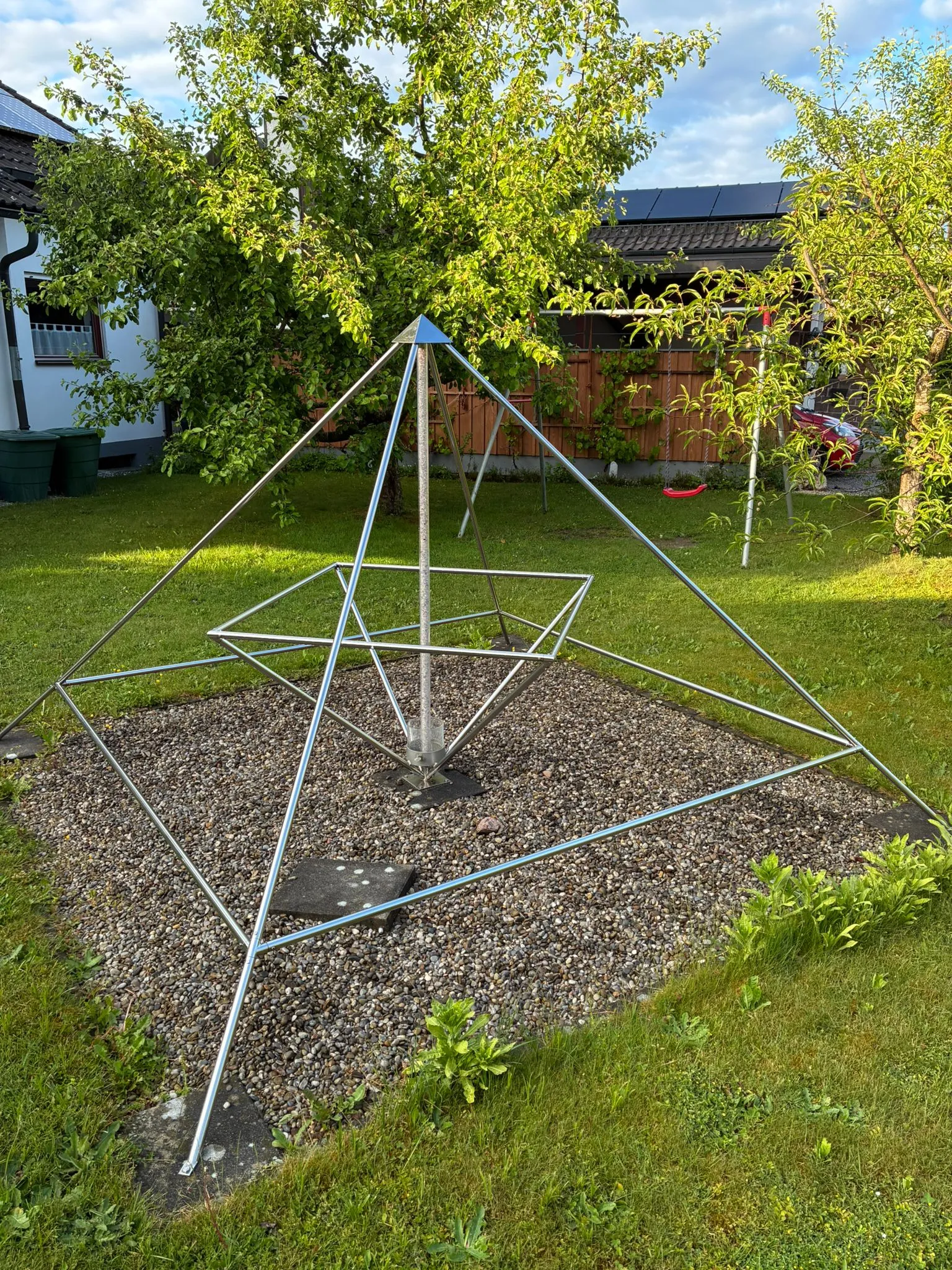 Große Energiepyramide Modell C als Edelstahl-Doppelpyramide auf einem Kiesbett im Garten; im Hintergrund ein Wohnhaus mit Solaranlage und eine Kinderschaukel.