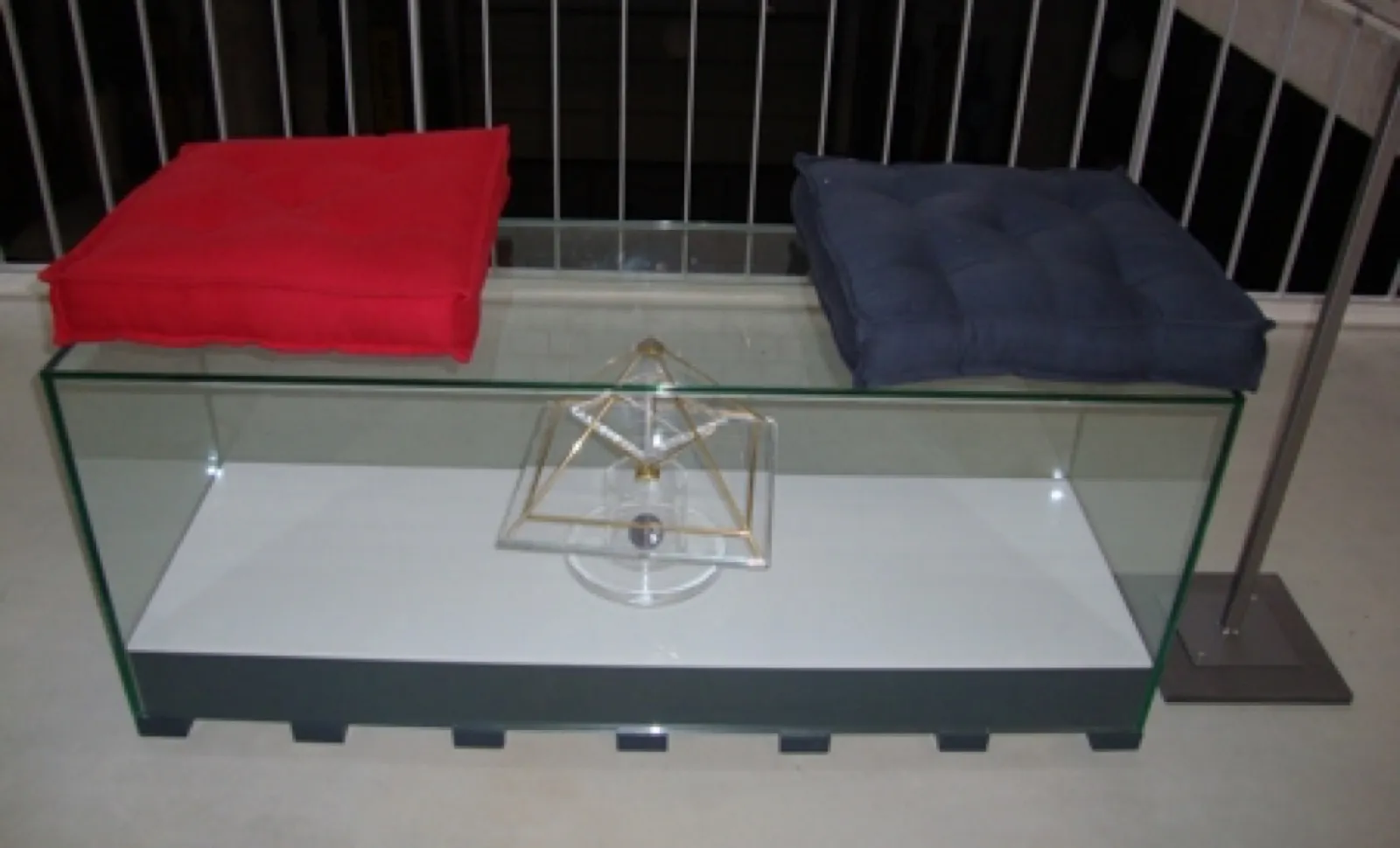 Eine Sitzbank aus Glas mit einem roten und einem dunkelblauen Sitzkissen; im Glaskörper darunter steht eine kleine vergoldete Energiepyramide mit Acrylglasachse, dahinter ein Geländer.