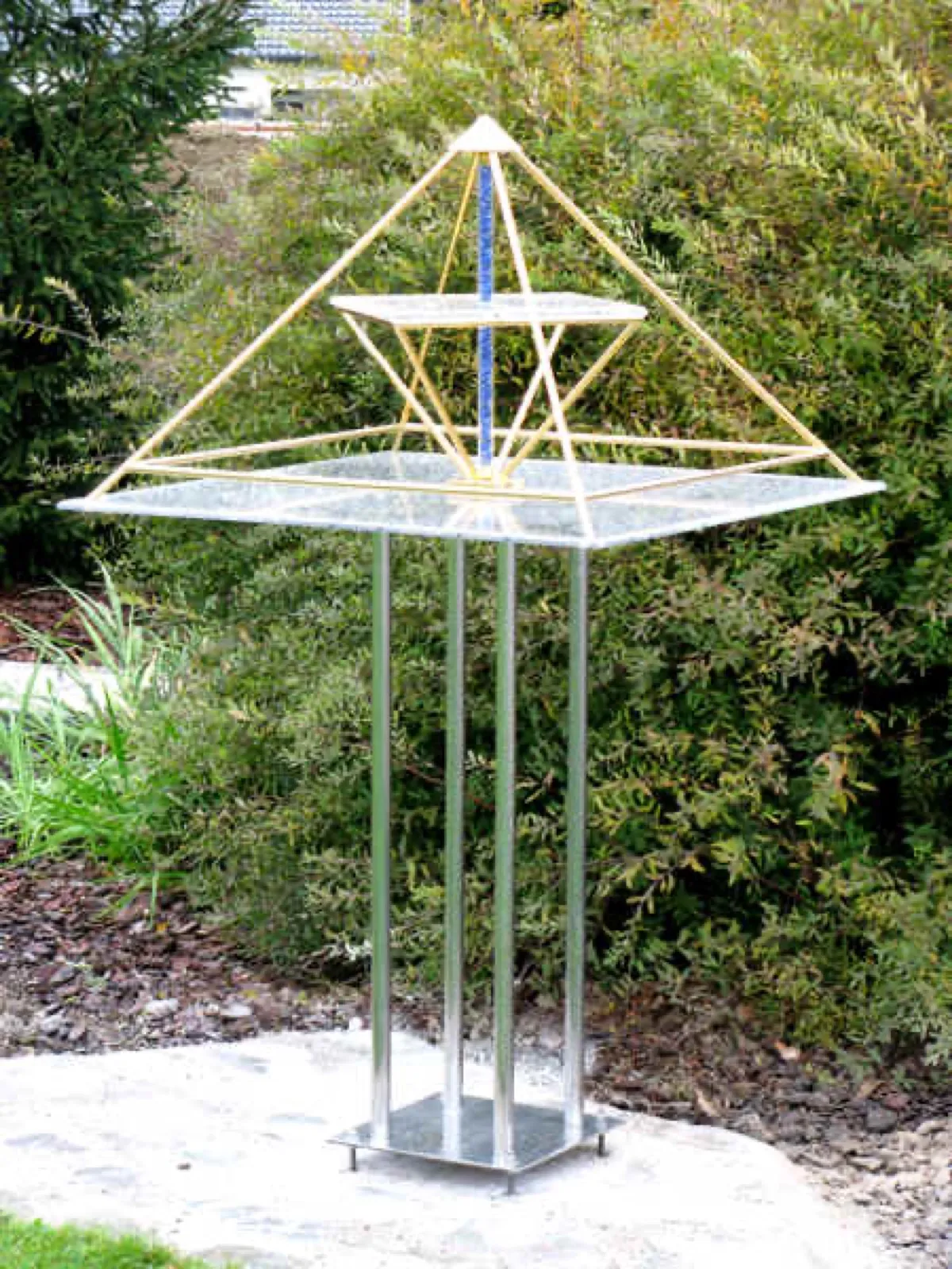 Eine goldfarbene offene Energiepyramide mit blau-weißer Mittelachse und Glasebenen steht auf einem hohen Gestell aus vier Edelstahlstützen auf einer Steinplatte im Garten. Dahinter dichte grüne Sträucher und ein Nadelbaum.