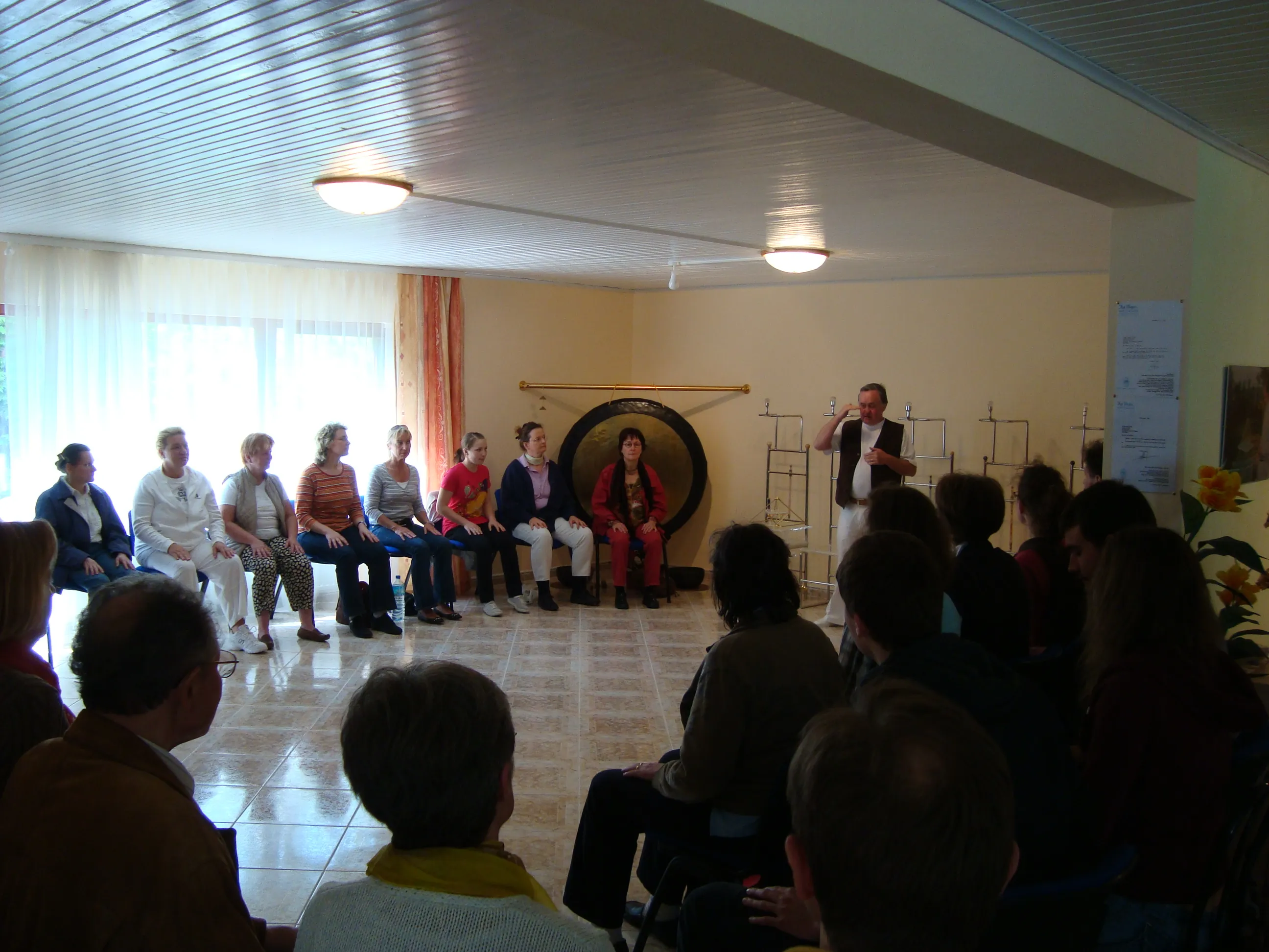 D. Harald Alke steht rechts und spricht bei einem Seminar im Kyborg Institut zu im Kreis sitzenden Teilnehmenden; im Hintergrund ein großer Gong und goldene Energiepyramiden-Modelle.