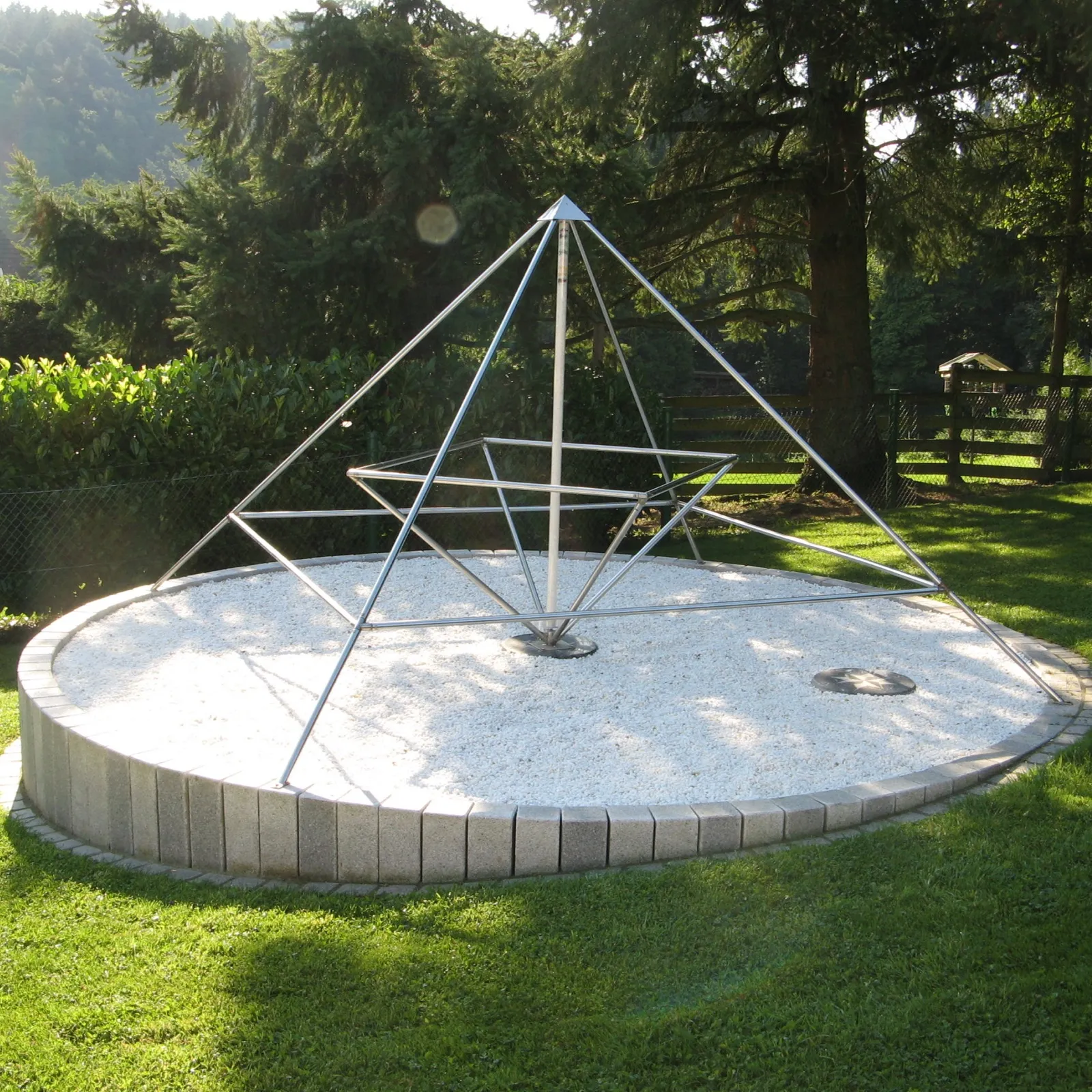 Offene Pyramide aus Edelstahlrohren mit innerem, verdrehtem Rahmen auf einem runden, weiß bekiesten Podest mit Betoneinfassung im sonnigen Garten; links oben neben der Spitze ein heller, runder Lichtfleck, dahinter Nadelbäume und ein Holzzaun.