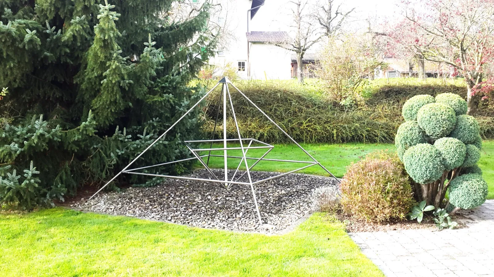 Offene Pyramide aus Edelstahlrohren mit innerem, verdrehtem Rahmen und mittiger Achse auf einem Kiesbeet in einem gepflegten Garten, zwischen einer großen Fichte und kugelig geschnittenem Formgehölz; dahinter ein weißes Haus.