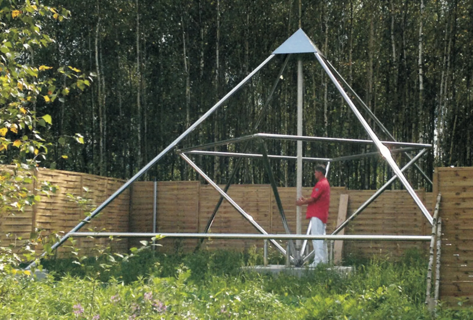 Ein Mann in rotem Poloshirt steht an der Mittelachse einer großen offenen Energiepyramide aus Edelstahlrohren, Modell F, in einem von einem Holzzaun umgebenen Garten; dahinter ein dichter Birkenwald.