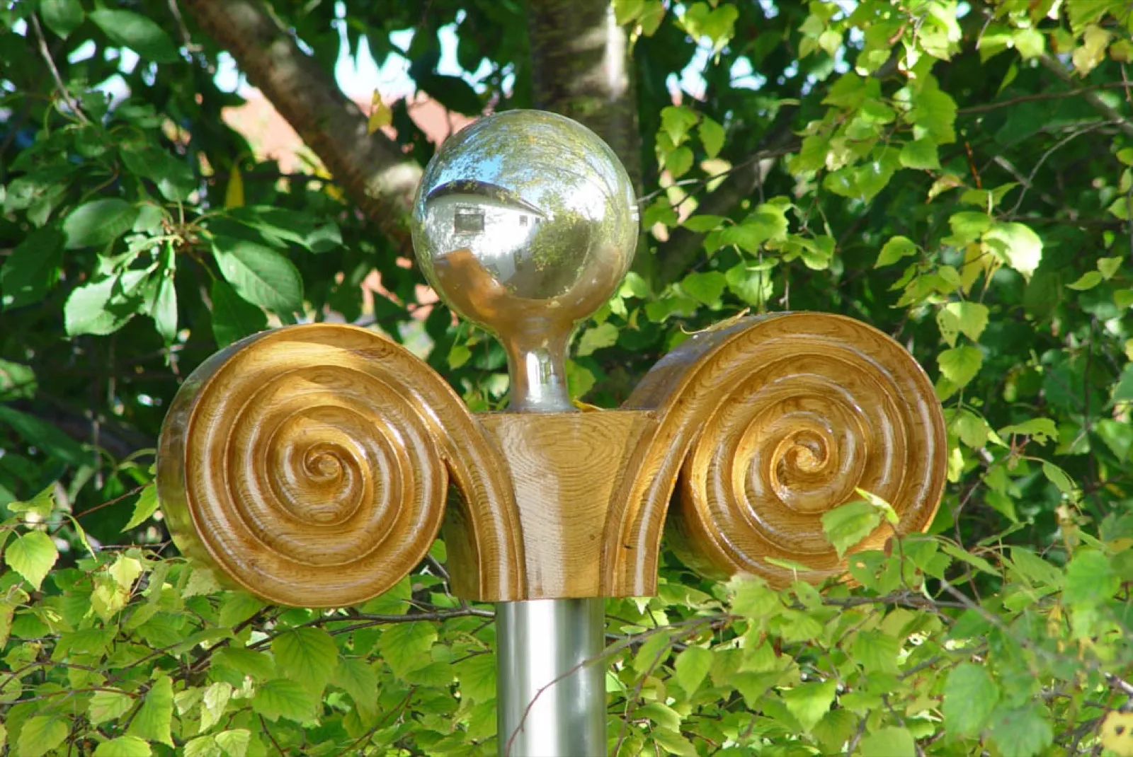 Holzkapitell einer Irminsul mit zwei geschnitzten Spiralvoluten und einer Spiegelkugel, in der sich ein Haus spiegelt, auf einem Edelstahlmast vor grünem Birkenlaub.