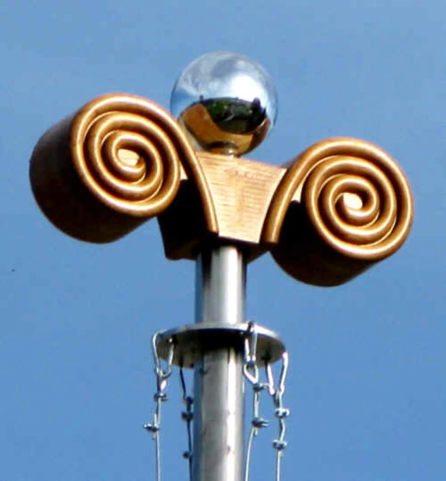 Spitze einer Irminsul vor blauem Himmel: goldfarbenes Kapitell mit zwei Spiralvoluten und aufgesetzter Spiegelkugel auf einem Edelstahlmast, darunter ein Ring mit herabhängenden Ketten.