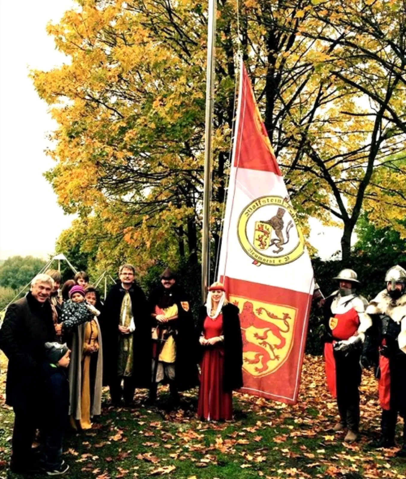 Eine Gruppe von Erwachsenen und Kindern, mehrere in mittelalterlichen Gewändern und zwei in Ritterrüstung, steht unter herbstlichen Bäumen neben einem Fahnenmast mit rot-weißer Fahne mit Löwenwappen. Links im Hintergrund die Spitze eines offenen Pyramidengestells.