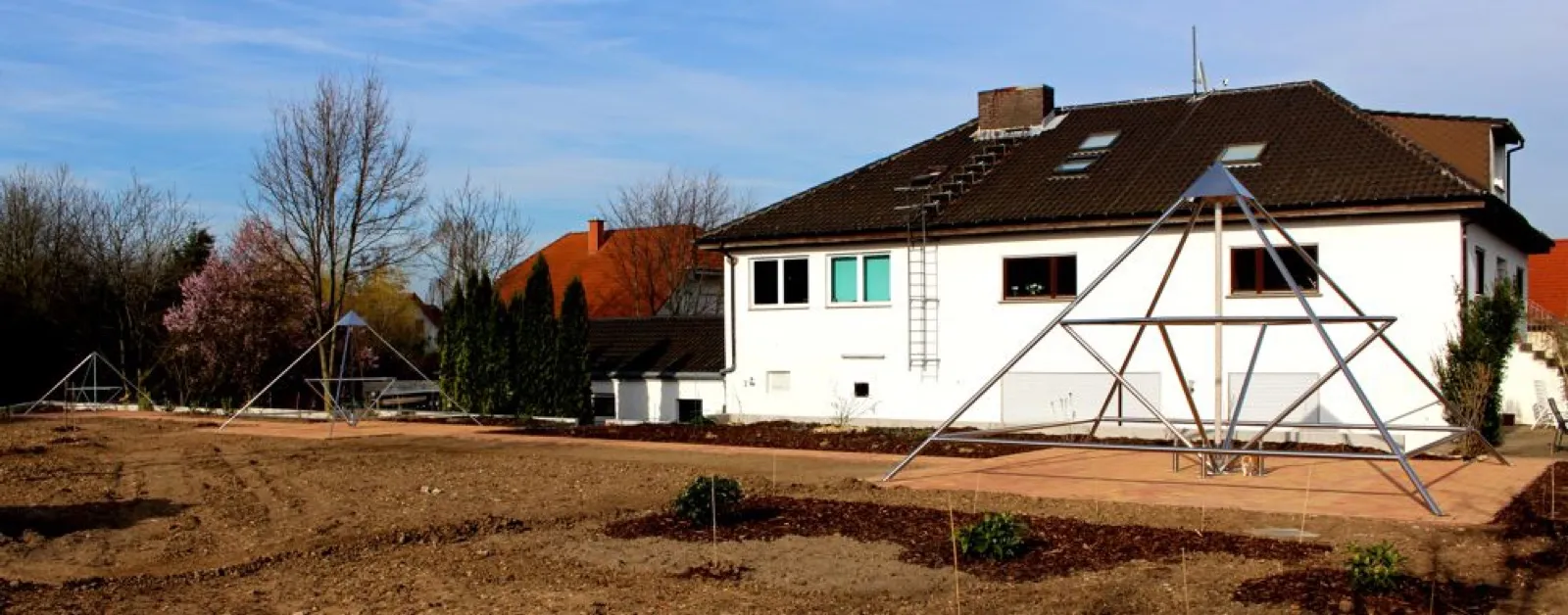 Neu angelegter Garten des Kyborg Instituts im Frühling: Vor dem weißen Institutsgebäude mit dunklem Dach steht eine große Energiepyramide aus Edelstahl auf einem gepflasterten Platz, links im Hintergrund zwei weitere Pyramiden neben einem blühenden Baum.