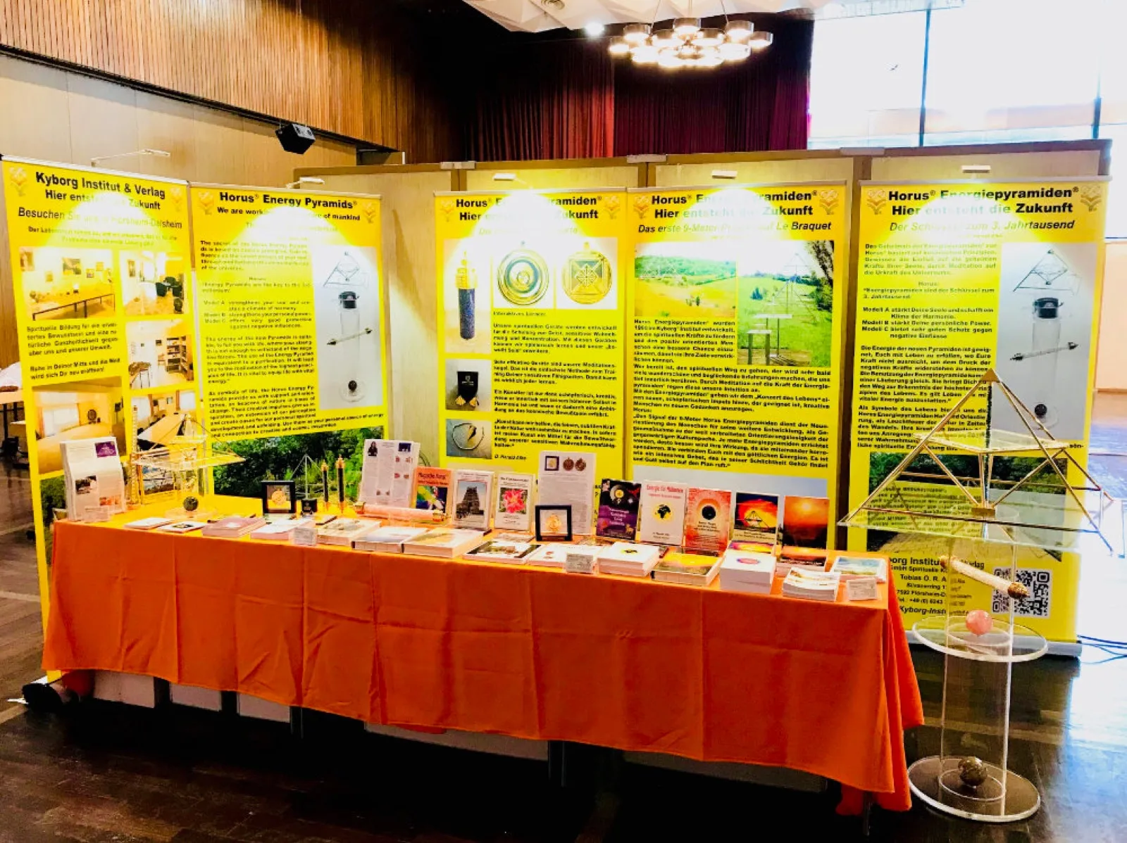 Messestand in einem Saal mit Holzvertäfelung und Kronleuchter: gelbe Roll-up-Plakate zu Kyborg Institut und Horus Energiepyramiden hinter einem orange gedeckten Tisch mit Büchern, Broschüren und kleinen Pyramidenobjekten. Rechts eine goldfarbene Pyramide über einem Acrylglas-Ständer mit Kugeln.