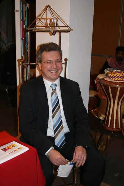 Markus Ferber im dunklen Anzug mit Brille sitzt lächelnd auf dem Horus Pharaonenstuhl, über dessen Sitz sich ein goldenes Pyramidengestell wölbt; im Hintergrund eine weiße Säule und bunte Trommeln am Messestand.