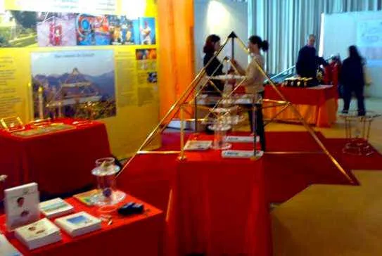Messestand mit rot gedeckten Tischen, einer großen goldenen Horus-Energiepyramide und kleineren Modellen, Kristallobjekten und Infotafeln; Personen im Hintergrund.