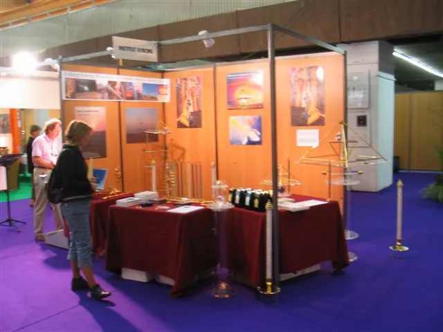 Messestand mit Holzwänden und Pyramiden-Postern, dunkelrot gedeckten Tischen mit goldenen Energiepyramiden und Kristallsäulen; Besucher auf violettem Messeteppich.