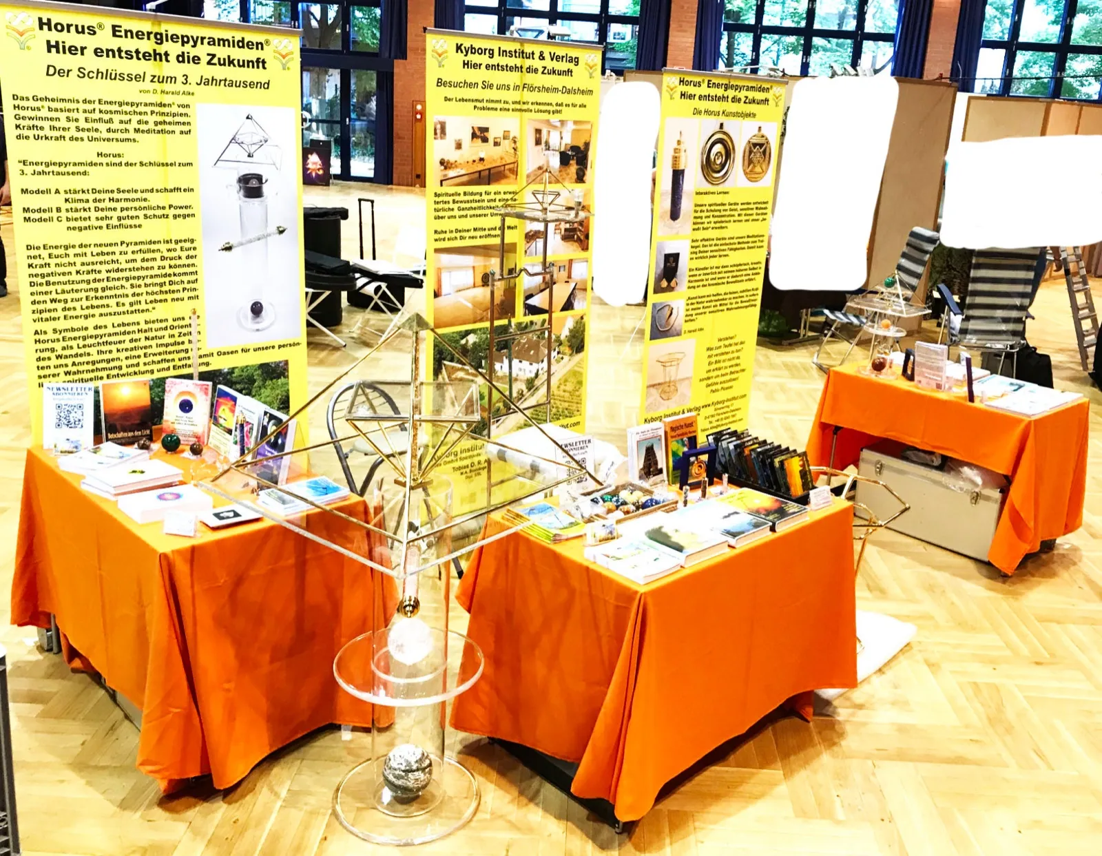 Messestand des Kyborg Instituts in einer Halle mit Parkettboden: Tische mit orangefarbenen Decken voller Bücher, Karten und Steinkugeln, davor eine goldfarbene Energiepyramide mit Acrylglasplatte auf einem Acrylglasständer mit Kristall- und Steinkugel; dahinter gelbe Roll-up-Banner.