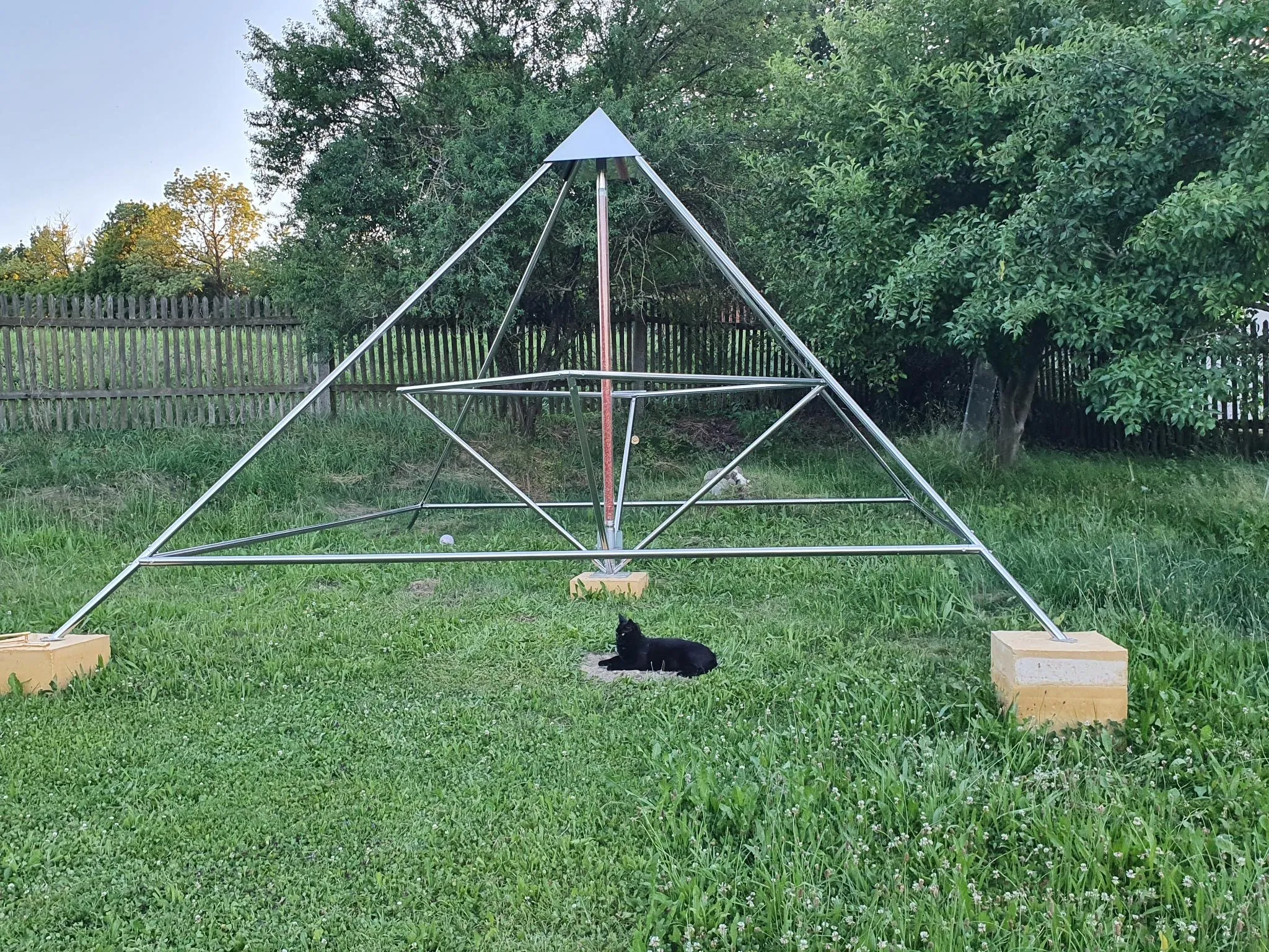Eine Horus Energiepyramide Modell D als silberne Metallrahmen-Doppelpyramide auf einer Rasenfläche in einem eingezäunten Garten; darunter liegt ein schwarzer Hund.