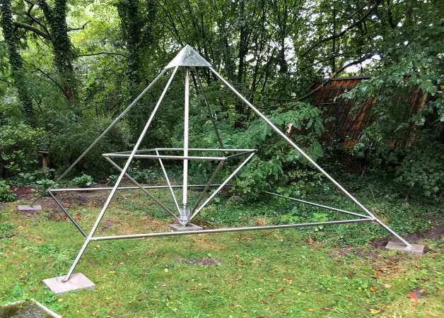 Eine Horus Energiepyramide Modell D als silberne Metallrahmen-Doppelpyramide steht in einem grünen Garten zwischen Bäumen.