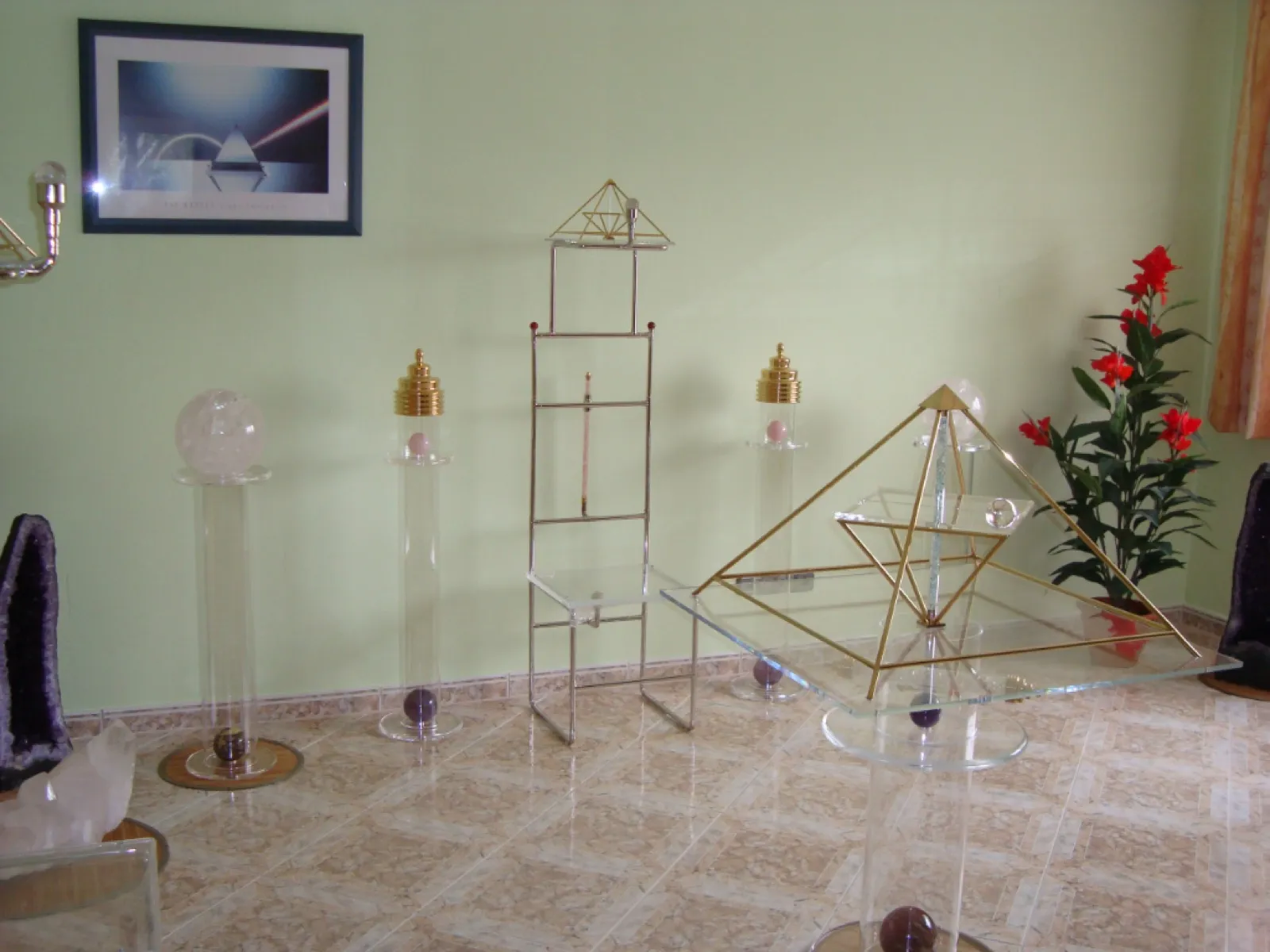 Raum mit hellgrüner Wand und Fliesenboden: in der Mitte ein schmaler Metallstuhl mit hoher Lehne und kleiner goldfarbener Pyramide obenauf, daneben Acrylglassäulen mit Bergkristallkugel und goldfarbenen Aufsätzen. Rechts vorne eine goldene Pyramide auf einem Glastisch, außerdem Amethystdrusen, ein Bergkristall, eine rot blühende Pflanze und ein gerahmtes Bild mit Pyramidenmotiv.