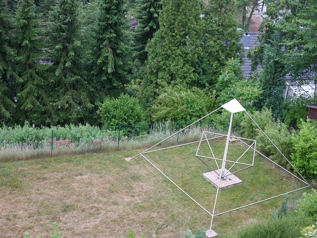 Blick von oben in einen Garten im Sommer: Rechts steht eine große offene Energiepyramide aus Edelstahlstäben mit innenliegendem Oktaeder und heller Spitze auf Steinplatten im Rasen. Links hinten, hinter einem Maschendrahtzaun im hohen Gras, ziehen ein Reh und ein Kitz vor dichten Nadelbäumen entlang.