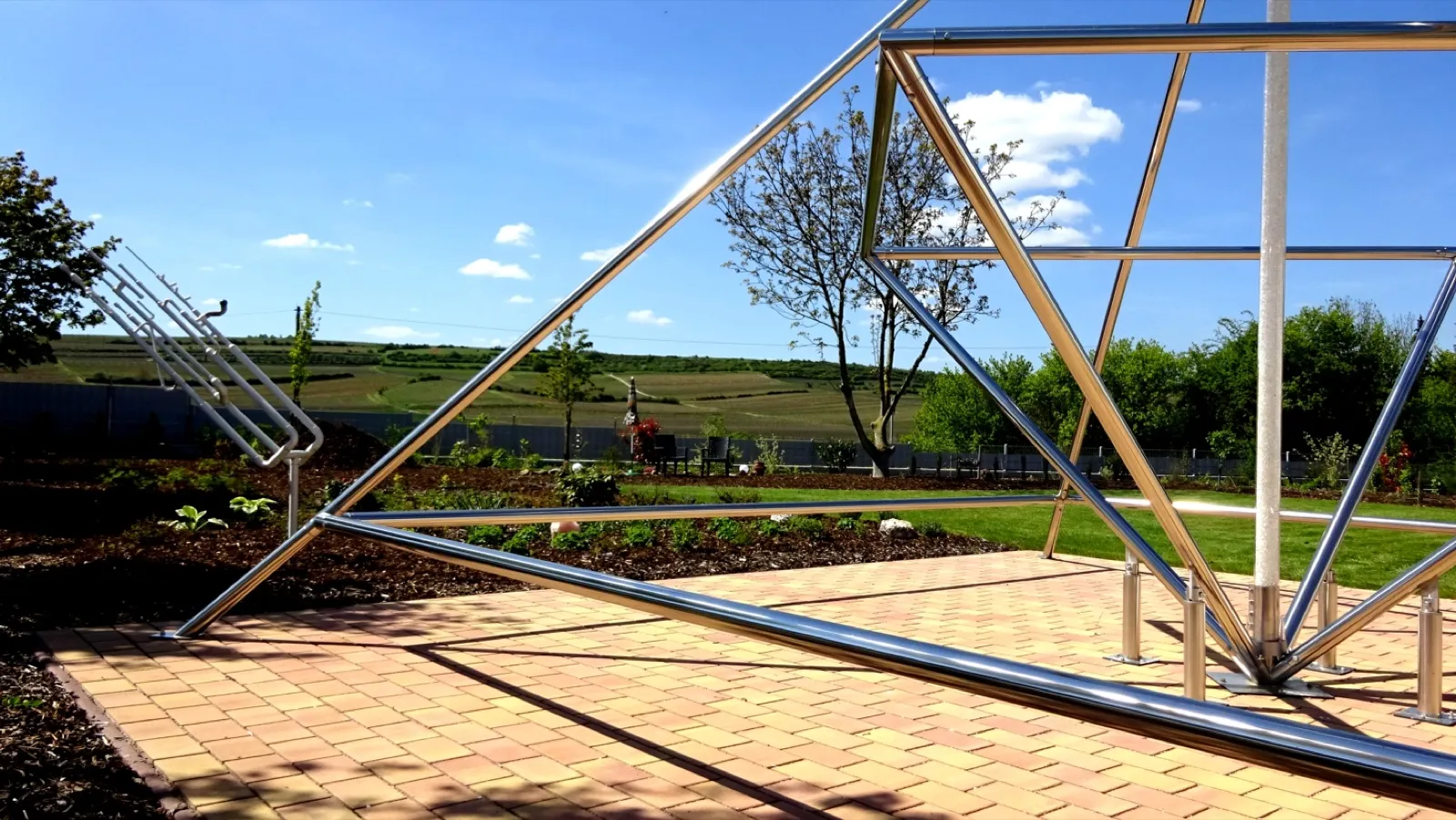Blick durch die glänzenden Edelstahlrohre einer großen Pyramide auf gepflasterter Fläche in einen angelegten Garten; links ein weißes Metallobjekt aus parallel gebogenen Rohren auf einem Ständer. Im Hintergrund Rasen, Beete, ein Baum, Gartenmöbel und sanft ansteigende Weinberge unter blauem Himmel.