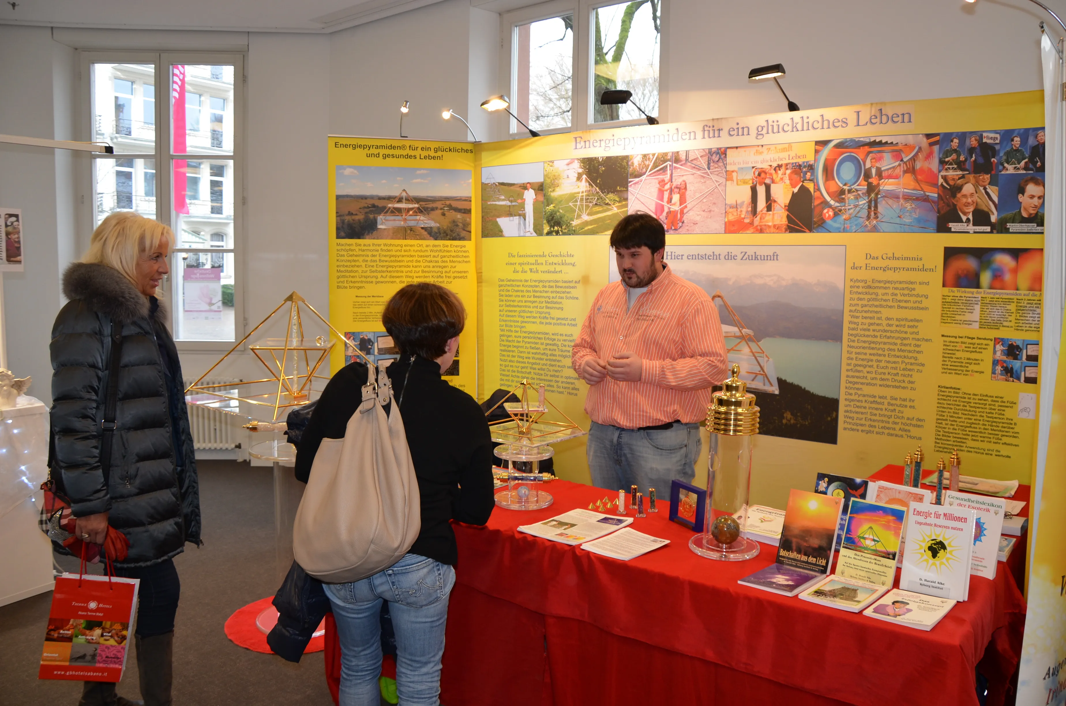 Tobias Alke im gestreiften Hemd im Gespräch mit zwei Besucherinnen am Energiepyramiden-Messestand; gelbe Infowände, goldene Pyramidenmodelle auf Acrylsäulen und ein roter Tisch mit Büchern und Edelstein-Stäben.