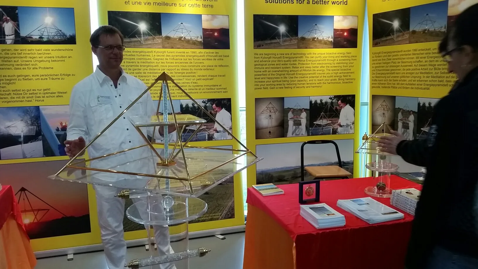Mats Björkman steht am Messestand des Kyborg Instituts neben einer goldenen Horus Energiepyramide auf einem Ständer; im Hintergrund mehrsprachige gelbe Rollup-Banner mit Produktinformationen.