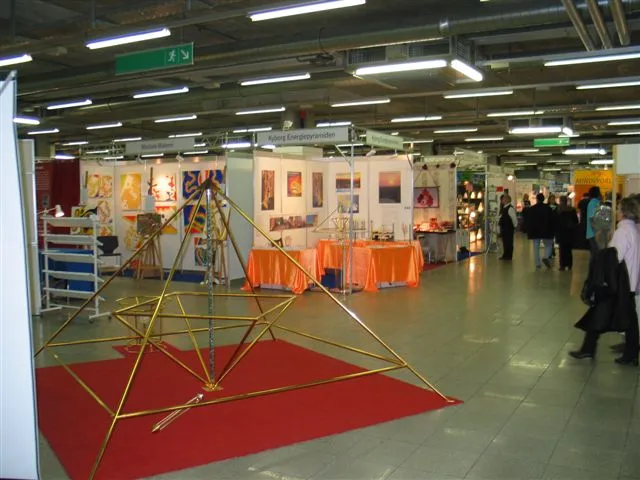 Messehalle mit dem Kyborg Energiepyramiden-Stand: im Vordergrund eine große goldene Horus Energiepyramide auf rotem Teppich, dahinter der weiße Standaufbau mit Fotos, orangefarbener Tischdecke und Dekorationselementen sowie weitere Stände und Besucher.