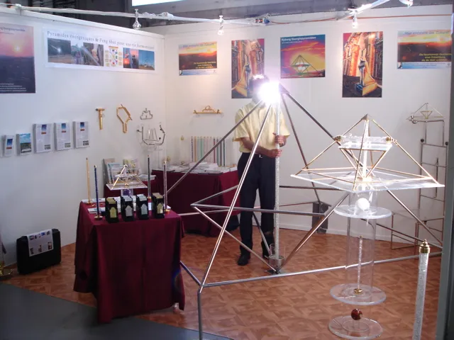 Messestand: ein Mann steht an einer großen goldenen Horus-Energiepyramide; dunkelrot gedeckte Tische mit Materialien, an der Wand französischsprachige Poster zu Energiepyramiden.