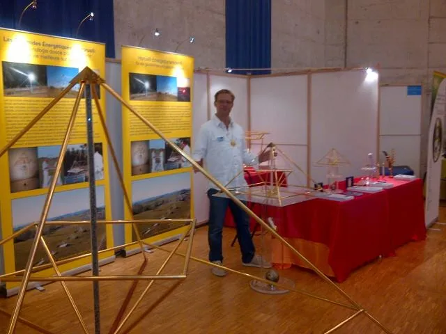 Mats Björkman steht an einem Messestand mit goldenen Horus Energiepyramiden verschiedener Größen sowie einem rot bedeckten Ausstellungstisch mit Modellen, im Hintergrund gelbe Infotafeln des Kyborg Instituts.