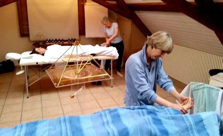 Blick in einen Seminarraum im Dachgeschoss: zwei Massageliegen mit liegenden Personen, zwei Frauen arbeiten an ihnen, in der Mitte eine goldene Horus Energiepyramide aus Metall.
