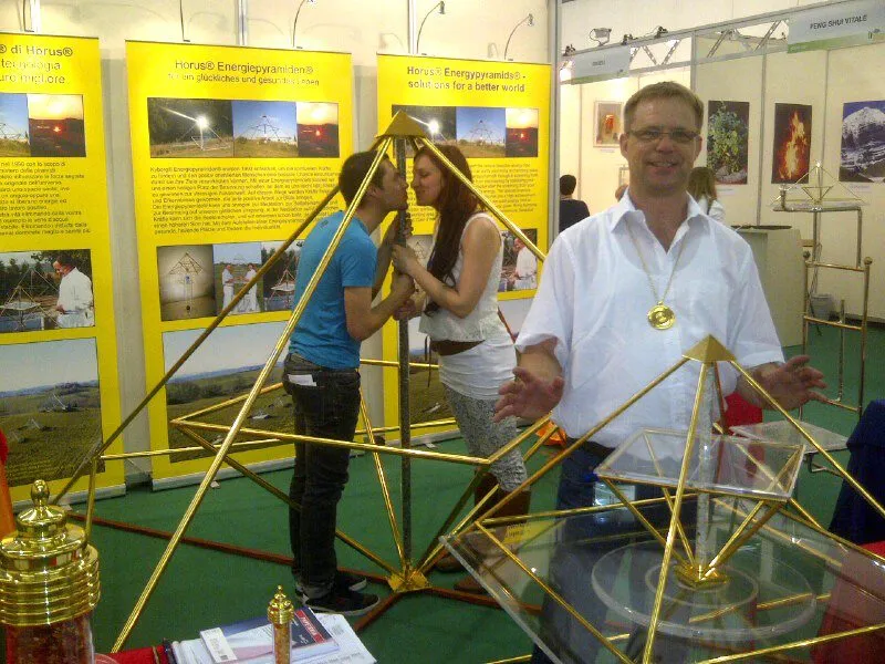 Mats Björkman am Horus-Energiepyramiden-Messestand mit goldenen Pyramidenmodellen im Vordergrund und gelben Ausstellungsbannern im Hintergrund.