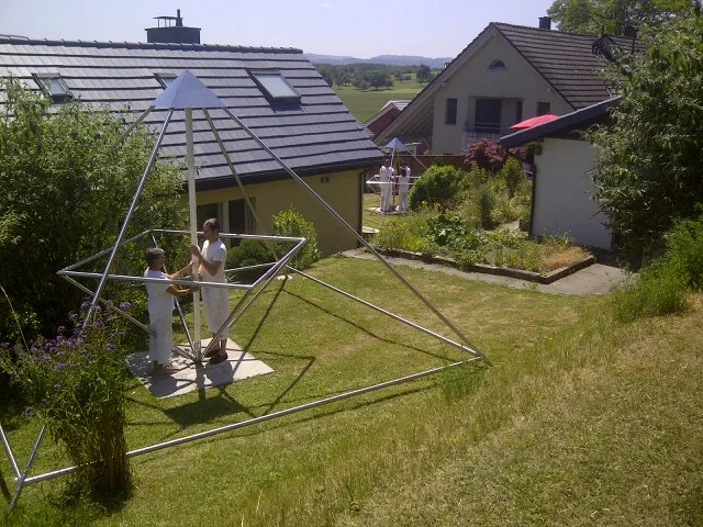 Zwei Personen stehen im Inneren einer großen, aus Metallrohren gefertigten Horus Energiepyramide im Garten eines Hauses in ländlicher Umgebung.