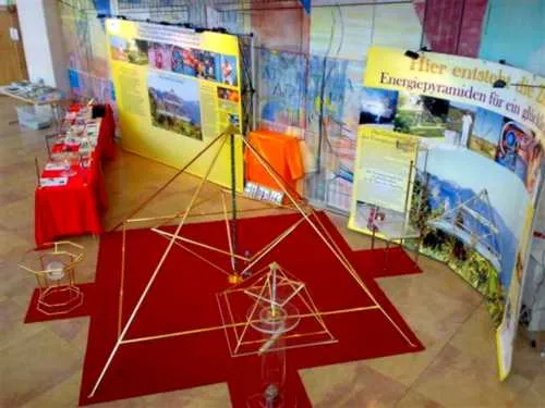 Ausstellungsfläche mit einer großen goldenen Horus-Energiepyramide auf einem roten, sternförmigen Teppich, gelben Infowänden und rot gedeckten Tischen.