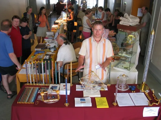 Ein junger Mann steht hinter einem mit rotem Tuch gedeckten Messestand, auf dem Horus Energiepyramiden, Stäbe und Informationsmaterial ausgestellt sind; im Hintergrund weitere Messestände und Besucher in einer hellen Messehalle.