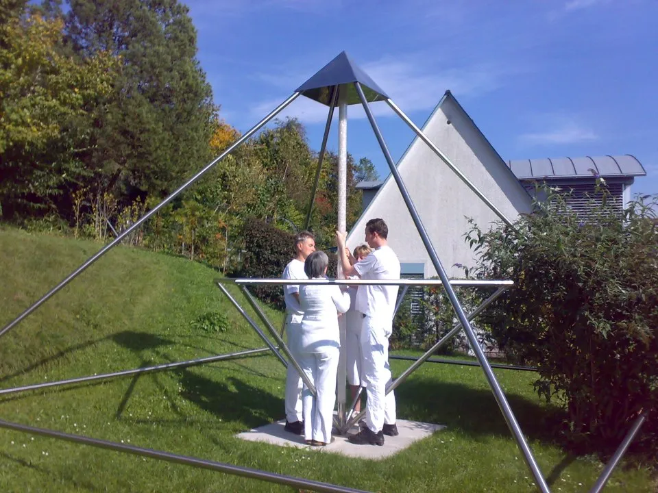 Drei Personen in weißer Kleidung stehen im Inneren einer großen Horus Energiepyramide aus Metallstreben im Freien, im Hintergrund Bäume und ein Gebäude bei sonnigem Wetter.