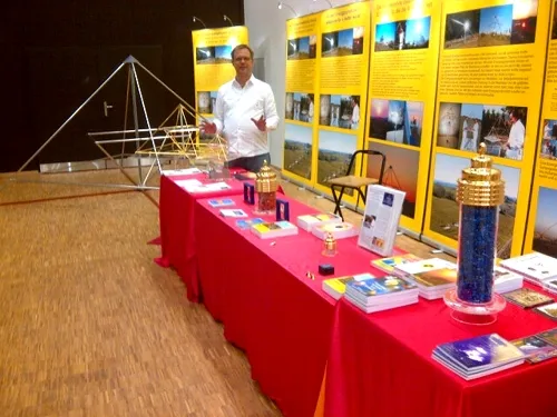 Mats Björkman steht hinter einem rot gedeckten Ausstellungstisch mit Horus Energiepyramiden-Modellen, Büchern und Informationsmaterial vor einer gelben Foto-Displaywand.