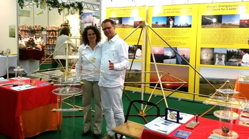 Eine Frau und ein Mann stehen an einem Messestand mit mehreren Horus Energiepyramiden-Modellen aus Metall vor einem gelben Hintergrund mit Werbebannern.