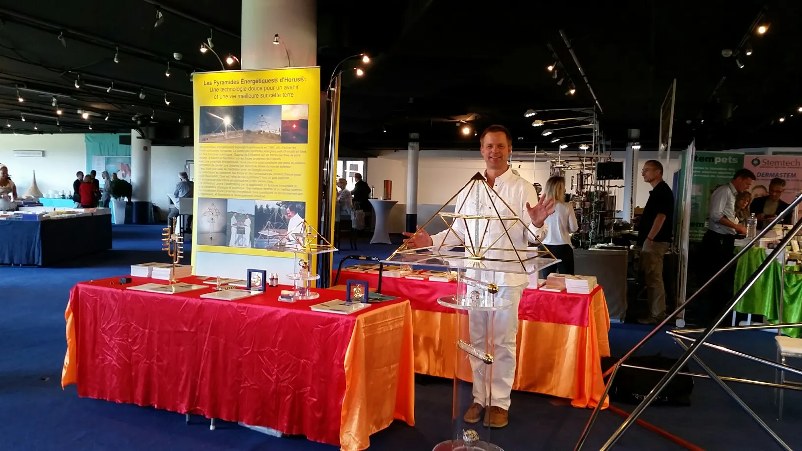 Mats Björkman steht hinter einem mit rotem und orangefarbenem Stoff dekorierten Messestand und präsentiert mehrere Horus Energiepyramiden unterschiedlicher Größen; im Hintergrund ein gelbes Ausstellungsbanner mit französischem Text sowie weitere Messestände in einem großen Hallenraum.