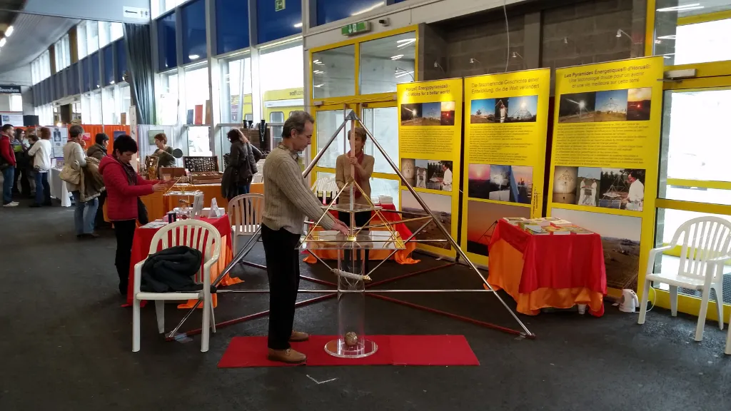 Mann steht vor einer großen Horus Energiepyramide aus Metall auf einem roten Teppich am Messestand des Kyborg Instituts; im Hintergrund gelbe Ausstellungstafeln mit Fotos und weiteren Besuchern.