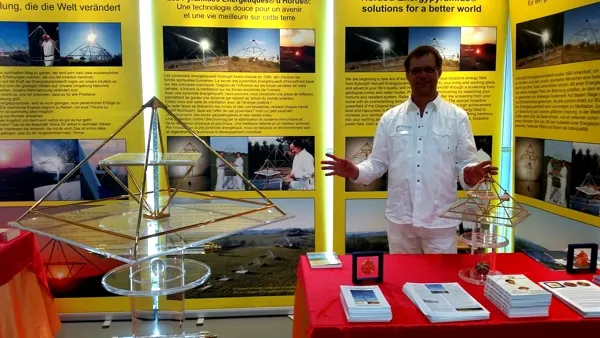Mats Björkman steht an einem gelb gestalteten Messestand mit zwei Horus-Energiepyramiden-Modellen aus Metall auf einem roten Tisch sowie Büchern und mehrsprachigen Ausstellungsplakaten im Hintergrund.