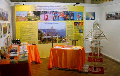 Messestand des Kyborg Instituts auf der Lebenskraft-Messe in Zürich 2008: orange drapierte Tische mit Broschüren und Flyern, im Hintergrund gelbe Informationstafeln, rechts ein freistehendes Horus-Energiepyramiden-Modell aus Metall und Acrylglas.