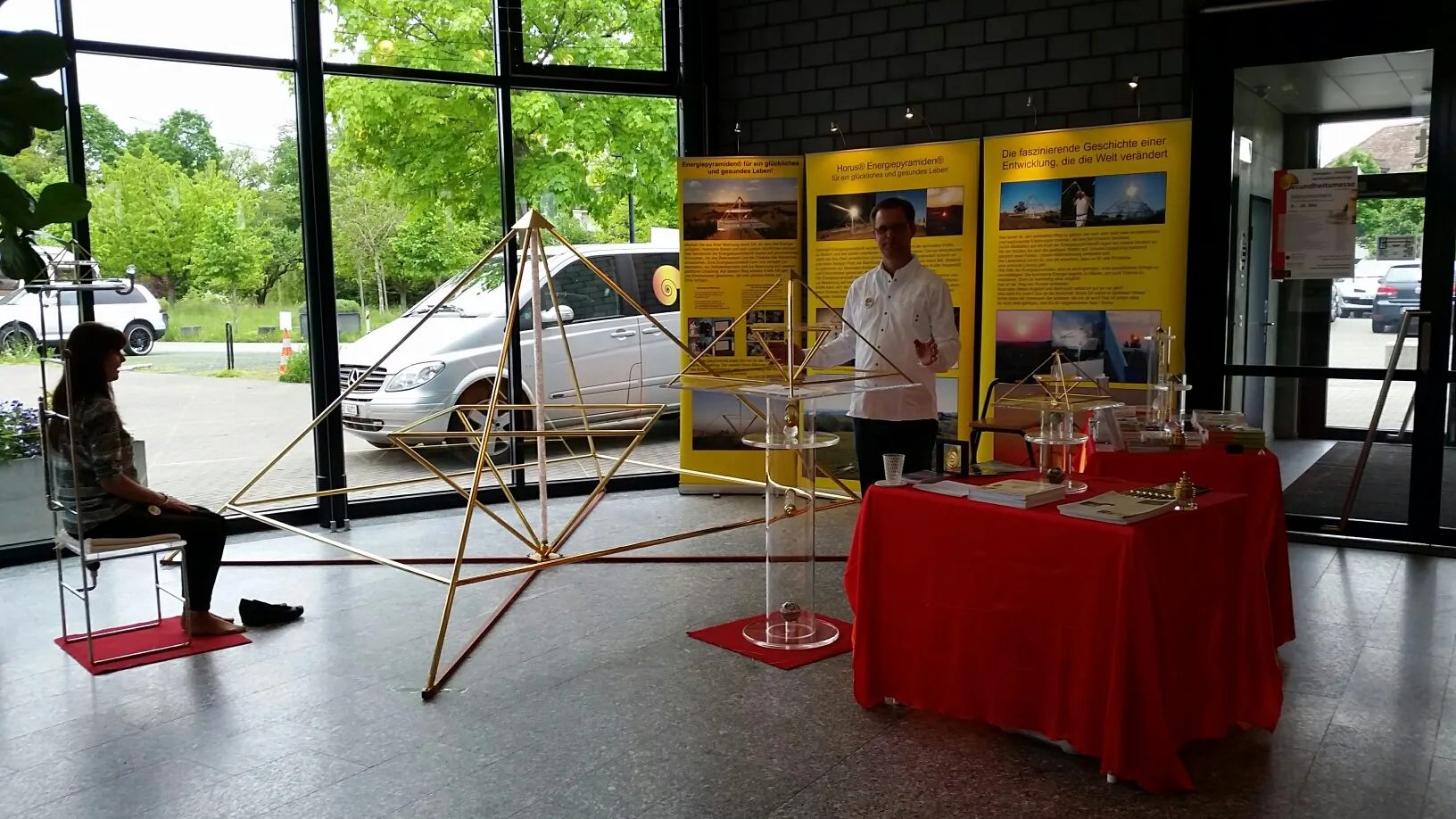 Messestand des Kyborg Instituts in einer Eingangshalle: Im Vordergrund eine große goldene Horus Energiepyramide, links sitzt eine Frau auf einem Stuhl, rechts steht Mats Björkman neben einem Stehtisch; im Hintergrund gelbe Roll-ups mit Pyramiden-Motiven.